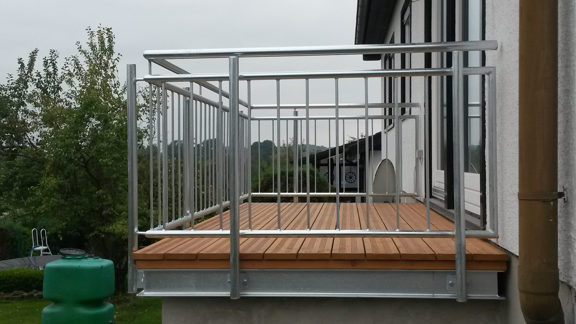 Kleiner Anbaubalkon aus feuerverzinktem Stahl mit Holzdielengang und vertikalem Stabgeländer an einer weißen Hauswand