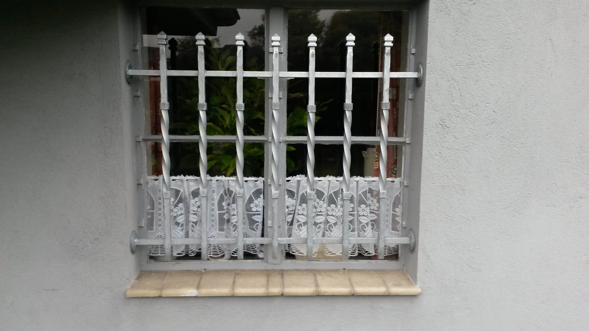 Hellgraues Fenstergitter mit gedrehten Stäben und Zierspitzen in der Fensterlaibung