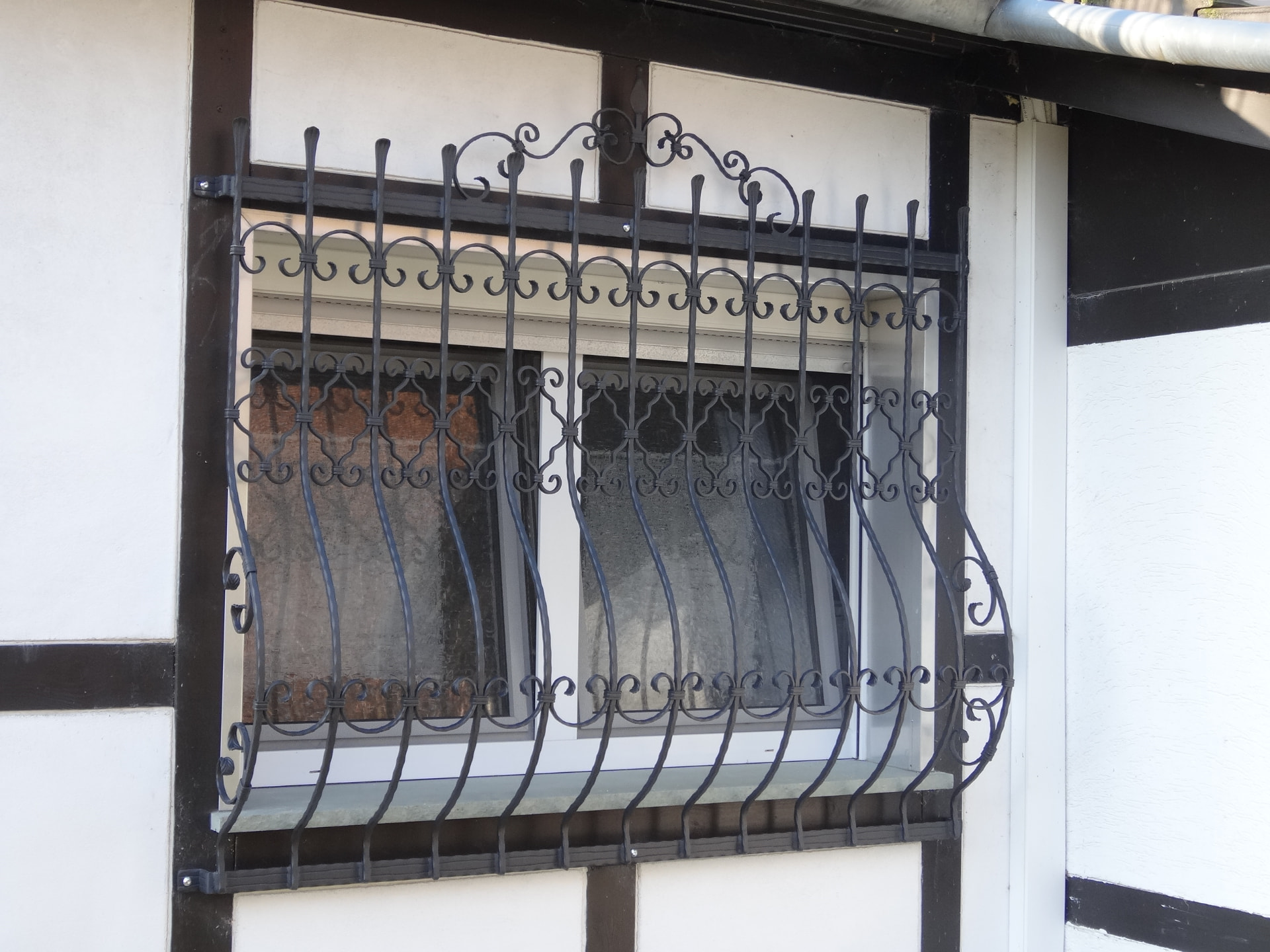 Schwarzes Schmiedeeisen-Fenstergitter mit S-Schnörkeln an einem Fachwerkhaus